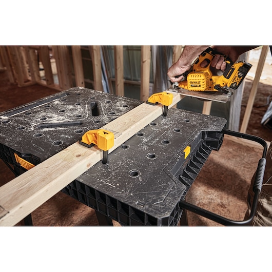 A DEWALT portable workbench with clamps securing a wooden plank, while a person uses a yellow DEWALT power tool to cut the wood in a construction setting.