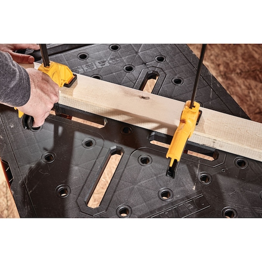 Close-up of a DEWALT workbench with a piece of wood clamped securely using two yellow clamps. A person's hands are adjusting one of the clamps. The black surface of the workbench has multiple holes and slots for versatility.