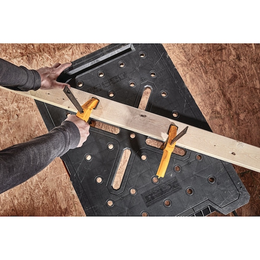 A person uses two yellow clamps to secure a wooden board onto a black DEWALT workbench.