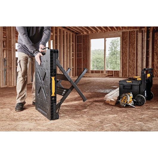 A person is unfolding a DEWALT portable workbench inside a building under construction. Next to the workbench are DEWALT toolboxes and a circular saw, placed on wooden boards.