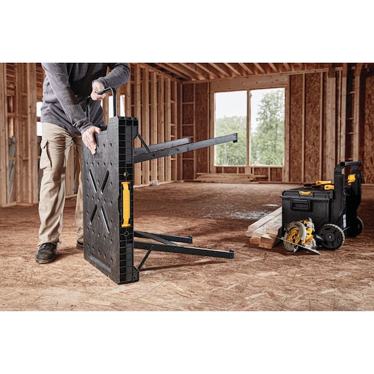 A person unfolds a black DEWALT portable workbench with yellow handle in an unfinished wooden construction site. Nearby are DEWALT storage boxes and tools, including a circular saw, placed on the floor beside some wooden planks.