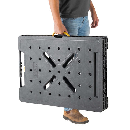 A person holding a DEWALT portable workbench with a black and yellow handle. The workbench is folded flat for easy carrying.