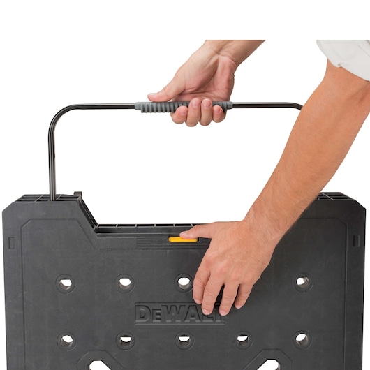 Close-up of two hands holding and carrying a DEWALT portable workbench with a black plastic body and a metal handle.