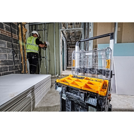 A DEWALT tool organizer box with yellow compartments containing screws and fasteners is open in the foreground on a construction site. In the background, a construction worker wearing a helmet and safety vest works on wiring inside a partially built wall. Sheets of drywall are stacked to the left.