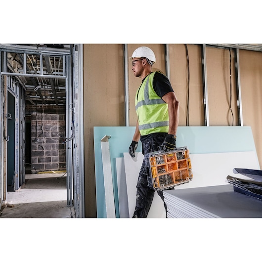 A construction worker wearing a reflective vest and helmet carries a DEWALT DWST82968-1 organizer in a building under construction.