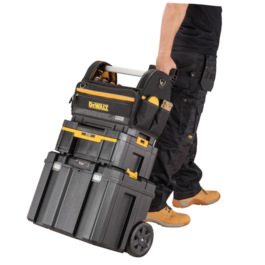 A person is pulling a stack of DEWALT portable toolboxes with wheels. The toolboxes are black with yellow accents and the DEWALT logo is visible on the top bag. The tool storage units are securely stacked, and the person is dressed in work pants and boots.