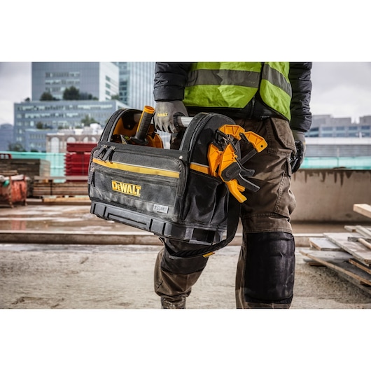 A person wearing work clothes and gloves is holding a DEWALT branded tool bag filled with various tools on a construction site. The tool bag has yellow accents and the DEWALT logo, with city buildings visible in the background.