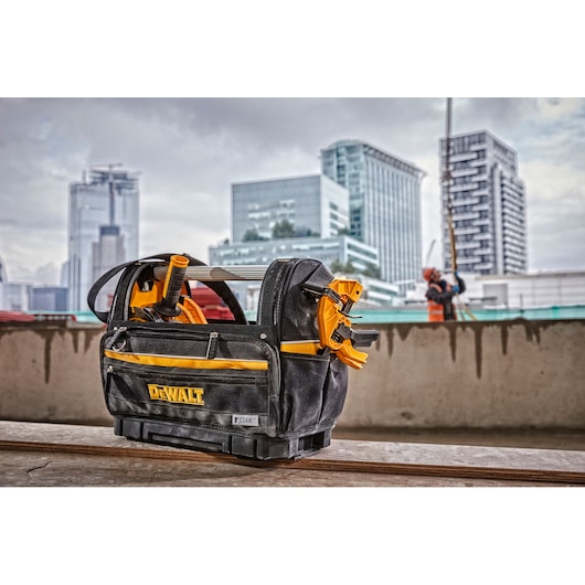 A DEWALT branded tool bag containing various tools, including clamps and a tape reel, is placed on a construction site with modern city buildings visible in the background.