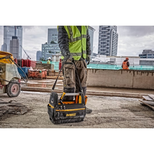 A DEWALT tool bag is being held by a person wearing work gloves, a green safety vest, and cargo pants, standing at a construction site with modern buildings in the background.