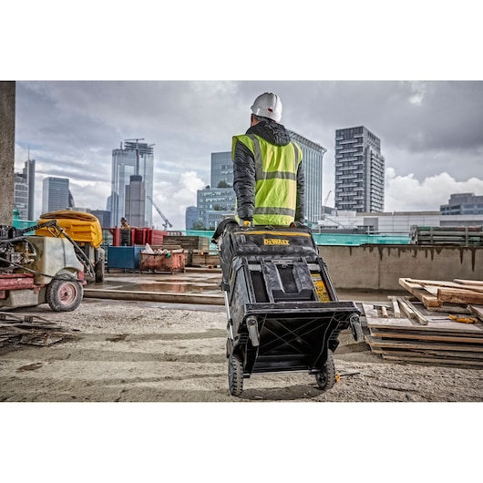 A person wearing a safety vest and hard hat on a construction site is pulling a large DEWALT rolling toolbox, surrounded by building materials and city skyscrapers in the background.