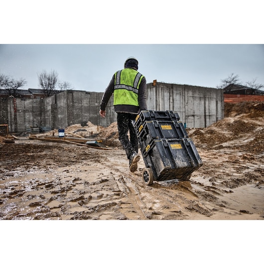 Worker pulls DEWALT toolboxes on wheels across a muddy construction site while wearing a safety vest and helmet.