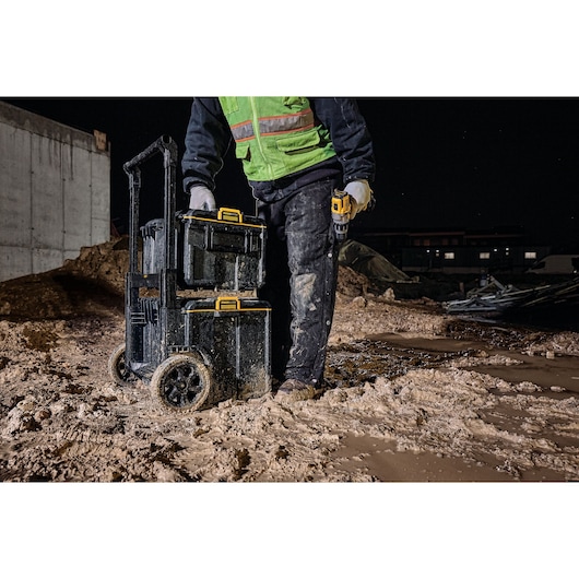 Person with DEWALT rolling tool box on muddy construction site at night, wearing safety gear and holding a drill.