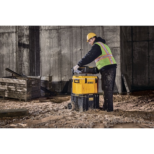Worker moves DEWALT wheeled toolbox on muddy construction site near wooden crates and concrete wall.