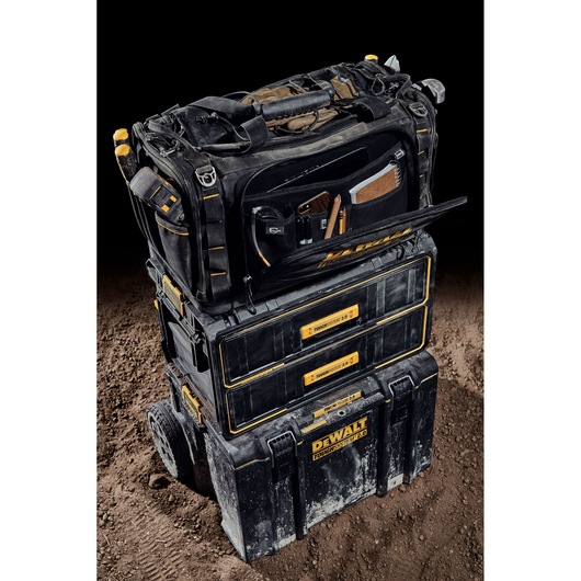 A DEWALT ToughSystem 2.0 tool storage setup is shown, consisting of three stacked black and yellow modular toolboxes. The top bag is open, revealing organized compartments with various tools and accessories. The setup is placed on a dirt surface with a black background.