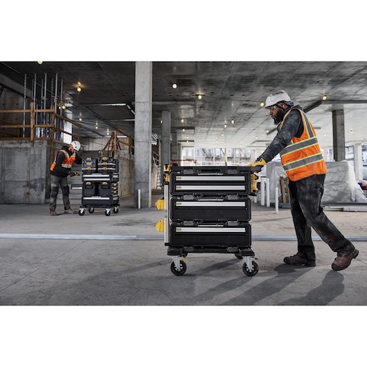 Two workers in safety gear and orange vests use DEWALT portable tool storage carts with multiple drawers on wheels inside a large construction site with concrete columns and overhead lights.